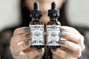 two hands reach out into the foreground with a person's face blurred in the background. The hands hold out two botany farms CBD oil jars with droppers as lids.