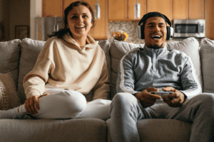 A couple sitting on a sofa in their room, the boy wearing headphones and holding a gaming joystick, both laughing and enjoying the game
