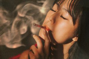 A woman with closed eyes holds a smoking cartridge to her mouth, exhaling smoke around her