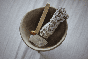A bundle of Sage, crystal, and palo santo wood in a pot on white surface, representing holistic elements for cleansing and positive energy