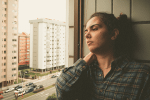 A tense-looking woman is sitting beside a window, looking out onto the street and There are some cars on the street and buildings on the other side