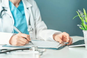 A doctor sitting in front of a desk, the torso from the neck down is visible with the arms resting on the table and taking notes on a note pad