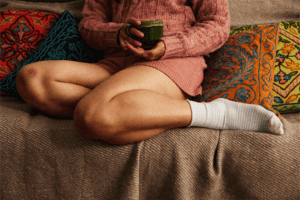 A person is sitting on a sofa, wearing a sweater, with a white sock on their leg, while holding a green cup in their hand