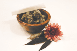 An arrangement featuring a small tub containing cannabis flowers, a rolled joint, a cannabis leaf, and a vibrant red flower placed beside the tub