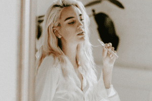A woman blow dab smoke as she lands against a wall with a vaporizer in hand.