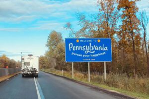 A scenic drive through Pennsylvania with a "Welcome to Pennsylvania" road sign, trees lining the road, a car with bicycles tied to the back, and a clear blue sky