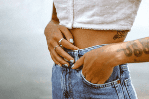 A woman in white top and blue jeans, left hand with tattoo in pocket, right hand on belly