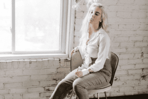 A woman sits on a chair next to a window and smokes from a Delta-8 TCH cartridge.